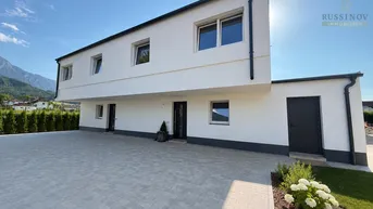 Modernes Einfamilienhaus mit weißer Fassade, Flachdach und gepflasterter Einfahrt vor Bergkulisse.