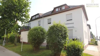 Weißes Mehrfamilienhaus mit mehreren Etagen und Dachgauben, umgeben von Grünflächen und Bäumen.