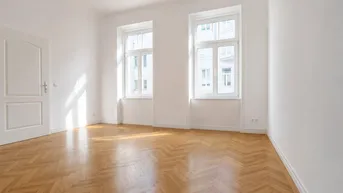 Helles, leeres Zimmer mit Fischgrätparkett und zwei großen Fenstern, das viel Tageslicht hereinlässt.