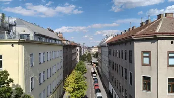 Blick über eine städtische Straße mit parkenden Autos und Gebäuden unter blauem Himmel.
