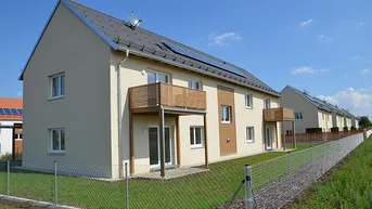Reihenhäuser mit moderner Architektur, Balkonen, Photovoltaikanlagen und grüner Umgebung unter blauem Himmel.