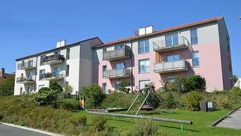 Mehrfamilienhaus mit Balkonen und einem kleinen Spielplatz im Vordergrund unter blauem Himmel.