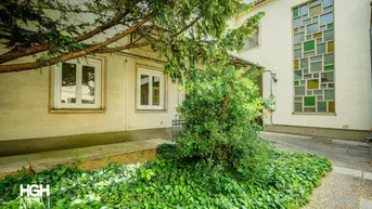 Außenansicht des Gebäudes mit dekorativen Fenstern, viel Grün und einem alten Baum im Vordergrund.