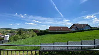 Panoramablick von einem Balkon auf eine weite grüne Landschaft mit Häusern und blauem Himmel.