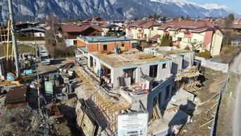 Luftaufnahme der Baustelle eines Neubaus mit Werbetafel und umliegender Wohnbebauung vor Bergkulisse.
