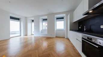 Heller offener Wohnbereich mit moderner Einbauküche, Fischgrätparkett und Zugang zu einem Balkon.