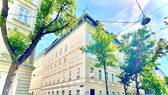 Klassisches Eckgebäude in einer sonnigen Stadtansicht mit grünen Bäumen und geparkten Autos.