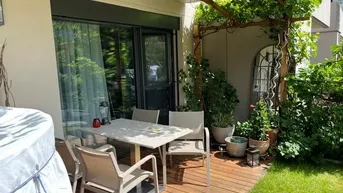 Sonnige Holzterrasse mit Esstisch, Stühlen und üppiger Begrünung unter einer Pergola.