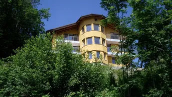 Gelbes Wohnhaus mit mehreren Balkonen, umgeben von dichtem Grün und hohen Bäumen.