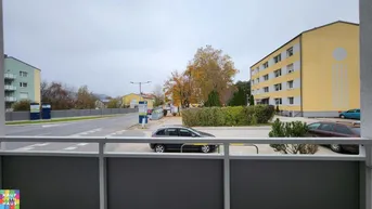 Blick vom Balkon auf eine Wohnstraße mit parkenden Autos, Bäumen und umliegenden Gebäuden an einem bewölkten Tag.