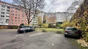 Außenansicht eines Wohnkomplexes mit Parkplätzen und Bäumen unter bewölktem Himmel.