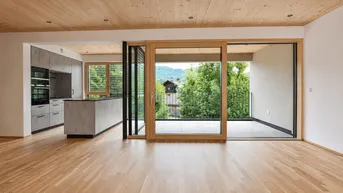 Heller offener Wohnbereich mit Holzboden, Holzdecke und integrierter Küche mit Zugang zum Balkon.