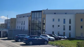 Modernes Mehrfamilienhaus mit Glasfassade und Außenparkplätzen unter blauem Himmel.