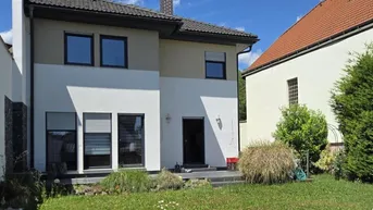 Modernes Einfamilienhaus mit weißer und beiger Fassade, umgeben von einem grünen Garten.