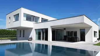 Moderne weiße Villa mit Flachdach, großem Swimmingpool und überdachter Terrasse unter blauem Himmel.