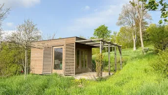 Kleines Holzhaus mit Terrasse in grüner Hügellandschaft unter blauem Himmel.