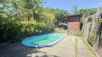 Kleiner Poolbereich im Garten mit Holzschuppen und üppiger Vegetation unter blauem Himmel.