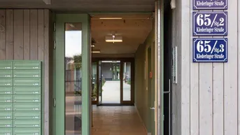 Moderner Eingangsbereich eines Mehrfamilienhauses mit heller Holzfassade, grüner Tür und Briefkästen, der in einen Flur führt.
