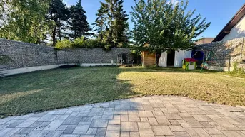 Großer Garten mit Rasenfläche, gepflasterter Terrasse und Spielbereich, umgeben von Gabionenmauern.