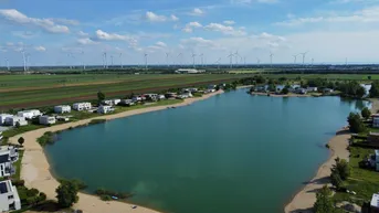 Luftaufnahme eines Sees mit umliegenden modernen Häusern und Windkraftanlagen im Hintergrund.