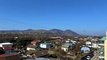 Weitläufiger Blick über eine Siedlung mit grünen Hügeln und Bergen im Hintergrund unter blauem Himmel.