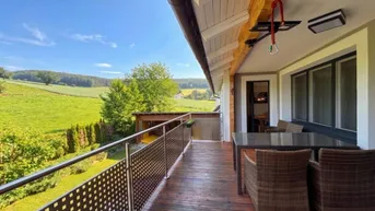 Geräumiger Balkon mit Holzboden, Sitzgelegenheiten und weitem Blick über grüne Felder und Hügel.
