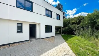 Moderne weiße Fassade eines Neubaus mit dunklen Fenstern und gepflastertem Eingangsbereich.