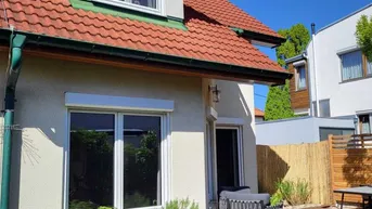 Sonnige Holzterrasse am Haus mit Gartenmöbeln und Blick auf den grünen Garten.