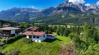 Luftaufnahme eines Hauses auf einem grünen Hügel mit majestätischer Bergkulisse im Hintergrund.