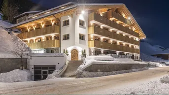 Modernes Gebäude im alpinen Stil, umgeben von Schnee, mit beleuchteten Balkonen in der Abenddämmerung.