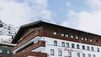 Ansicht des Hotels Kirlana mit traditioneller Holzfassade und schneebedeckten Bergen im Hintergrund.