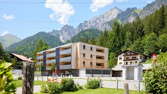 Modernes Gebäude mit Holzfassade und Balkonen, umgeben von grünen Bergen und Bäumen.
