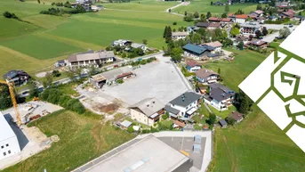 Luftaufnahme einer ländlichen Siedlung mit Wohnhäusern, grünen Feldern und einem See im Hintergrund.