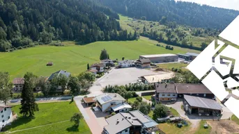 Luftaufnahme eines ländlichen Gebiets mit Wohnhäusern, grünen Wiesen und einem Wald im Hintergrund.