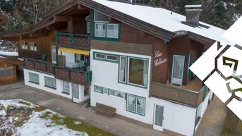 Traditionelles Alpenhaus mit mehreren Holzbalconen, schneebedecktem Dach und umgeben von Wald.