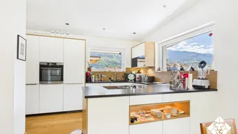 Moderne Küche mit weißen Schränken, Holzakzenten und dunkler Arbeitsplatte. Große Fenster bieten einen beeindruckenden Bergblick.
