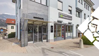 Fassade eines modernen Geschäftsgebäudes mit großem Eingangsbereich und Schaufenstern, umgeben von Bergen.