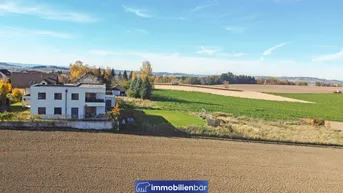 Weitläufige Landschaft mit Feldern und einem modernen Wohnhaus in der Ferne unter blauem Himmel.