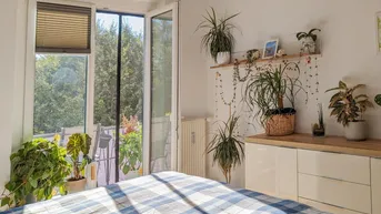 Helles Schlafzimmer mit Zugang zum Balkon, Holzboden und vielen Pflanzen.