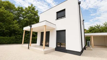 Modernes Einfamilienhaus mit weißer Fassade, dunklen Rollläden und Kiesauffahrt unter blauem Himmel.