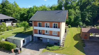 Mehrstöckiges Wohnhaus mit Balkonen, Garage und großem Garten, umgeben von viel Grün und Bäumen.