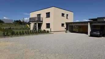 Modernes Einfamilienhaus mit beiger Fassade, Flachdach, Balkon und großem Kiesparkplatz mit Carport.