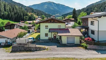 Haus in alpiner Umgebung mit Bergblick und umliegenden Wäldern unter blauem Himmel.