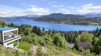 Panoramablick auf einen großen See mit umliegenden Wäldern und Bergen unter blauem Himmel.