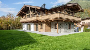 Traditionelles Holzchalet mit Steinfassade, großem Garten und Bergblick unter blauem Himmel.