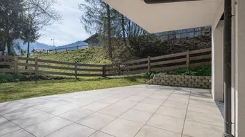 Große geflieste Terrasse mit angrenzender Rasenfläche und Holzzaun, umgeben von grüner Landschaft.
