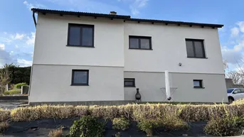 Modernes Einfamilienhaus mit heller Fassade und dunklen Fenstern unter blauem Himmel.
