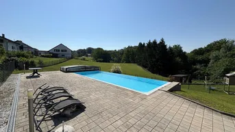 Großer Außenpool mit gepflasterter Liegefläche und weitem Blick ins Grüne unter blauem Himmel.
