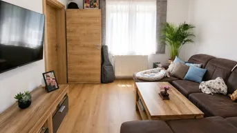 Helles Wohnzimmer mit braunem Ecksofa, Holzboden und einem großen Fernseher an der Wand.