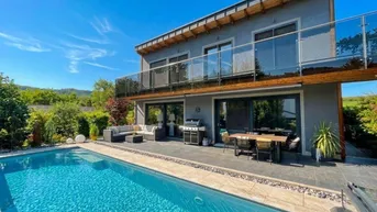Modernes Haus mit großem Pool, weitläufiger Terrasse und Balkon, umgeben von grüner Landschaft.